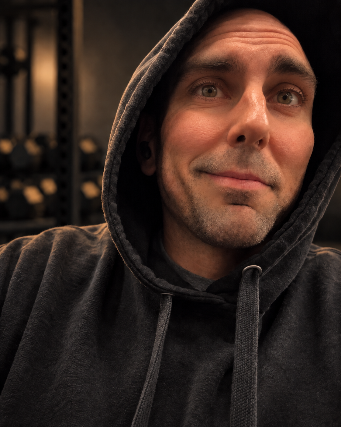 Alex in a charcoal hoodie looking toward the camera, against a softly lit dark fitness-studio background.